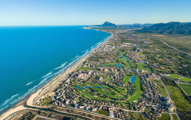 An aerial view of Oliva Nova, with blue skies, the sea and a long beach, green golf courses and mountains on the horizon.